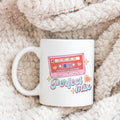A white ceramic mug with a retro typographical print featuring the words 'We are a Perfect Mix' and a cassette tape graphic, placed on a cozy, textured surface.