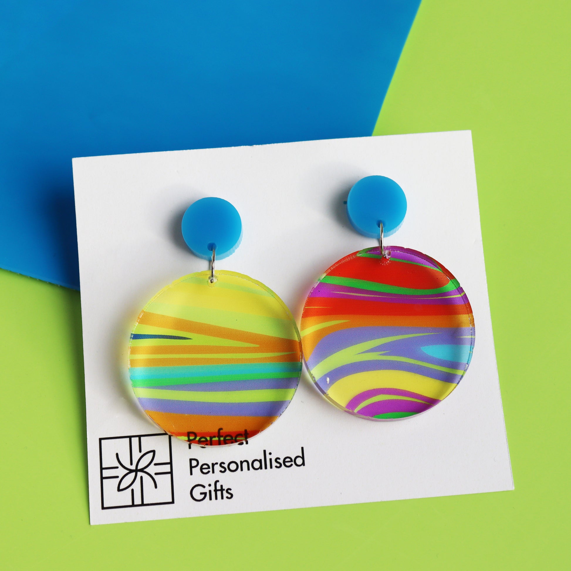 printed acrylic colourful mismatch earrings that have a round blue acrylic stud top and hanging printed acrylic circle hanging on a green backgrund