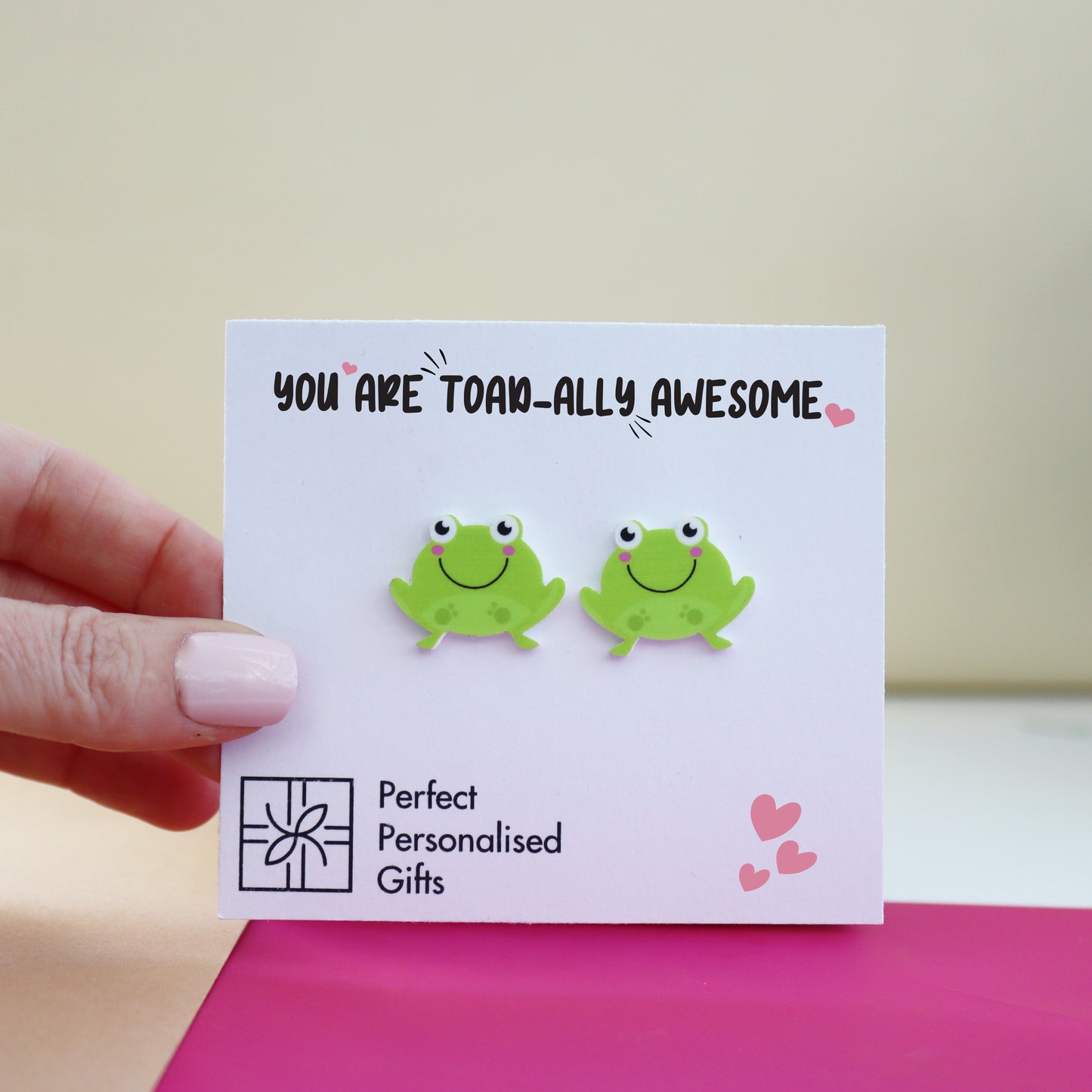 close up of cute you are toadally awesome frog acrylic earrings shown on pink and marble background