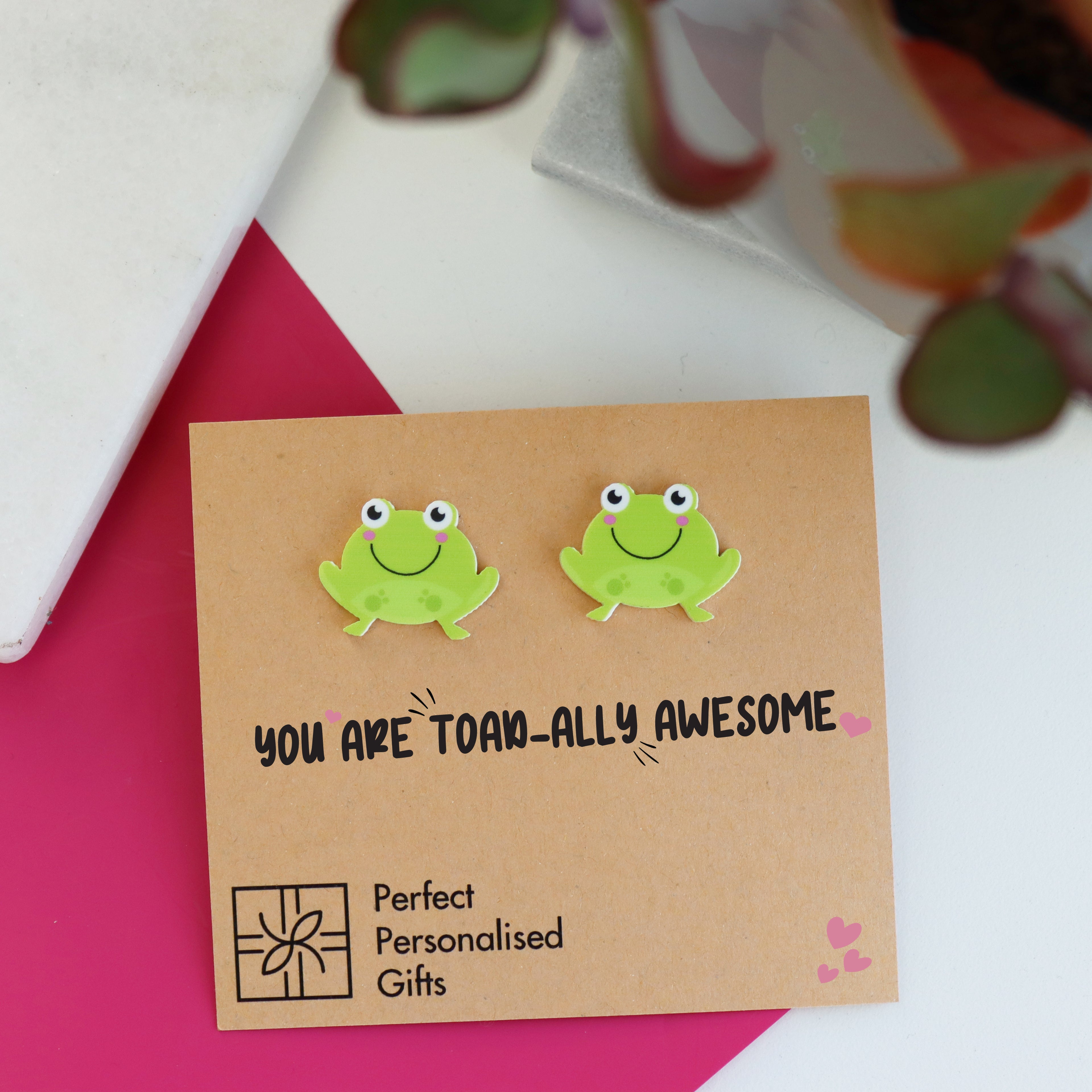 close up of cute you are toadally awesome frog acrylic earrings shown on pink and marble background